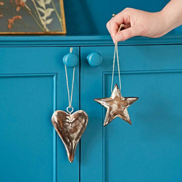 A hand holds **Mango Wood Heart and Star Decorations**, both hanging from strings on a blue cabinet.