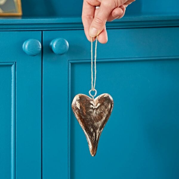 A hand holding a Mango Wood Heart and Star Decoration with a string loop, made from mango wood, against a blue wood cabinet with round knobs.
