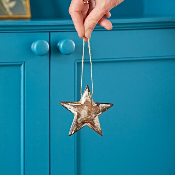 Hand holding a Mango Wood Heart and Star Decorations by a string in front of a blue cabinet with round knobs, perfect for adding to your star decorations collection.