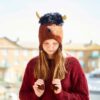 A girl wearing a Shaggy Bull Woollen Animal Hat and a red sweater looks down, with buildings and a bright sky in the background.