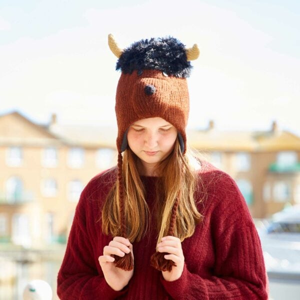 A girl wearing a Shaggy Bull Woollen Animal Hat and a red sweater looks down, with buildings and a bright sky in the background.