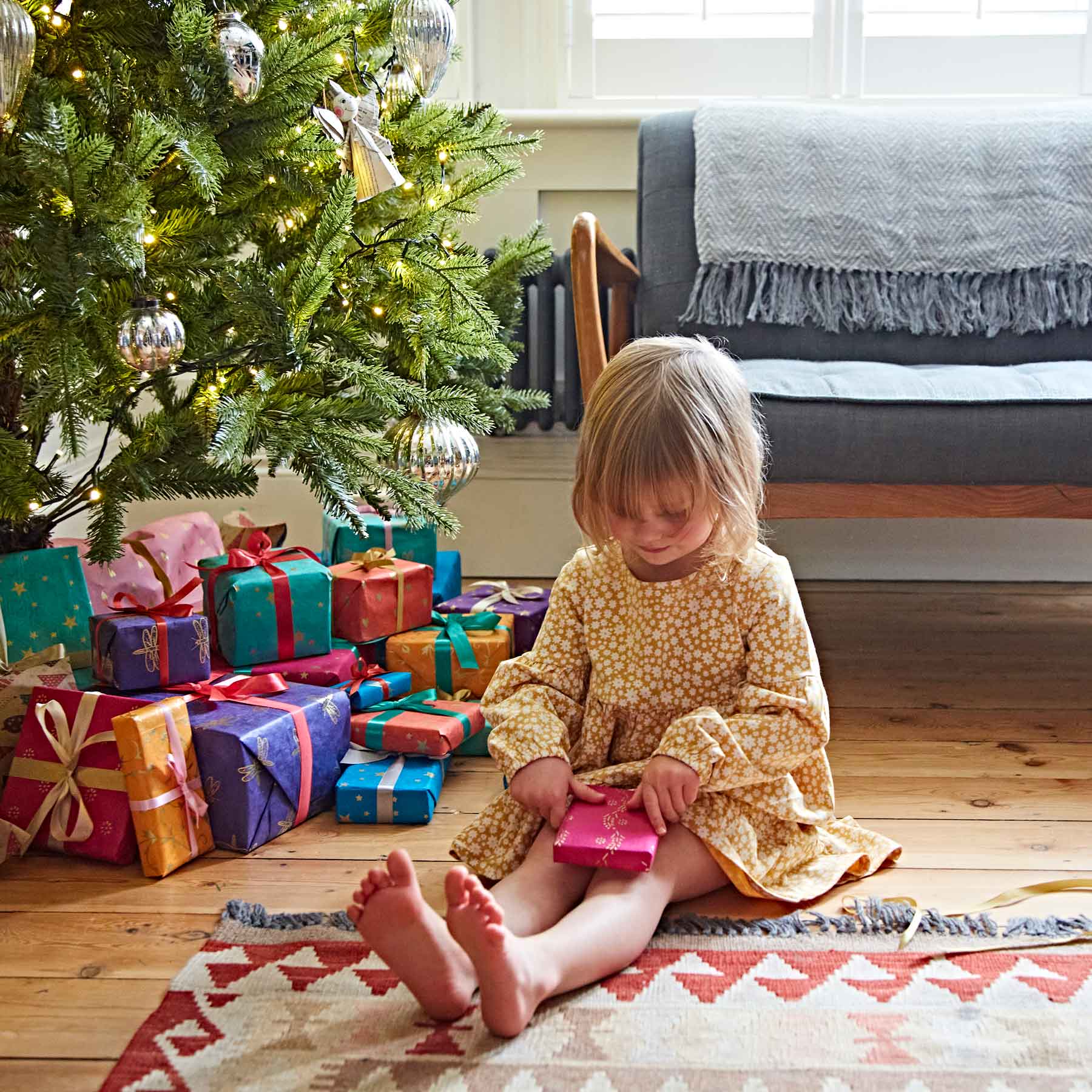 A young child in a yellow dress sits on the floor by a decorated Christmas tree, surrounded by multiple wrapped presents, holding a small gift—a heartwarming scene of Christmas joy.