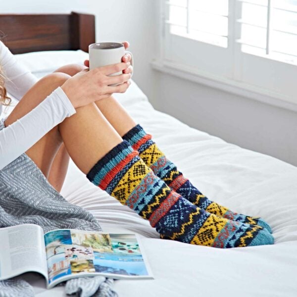 A woman sitting on a bed with hand knitted cosy socks on, holding a cup of coffee and looking at a book.
