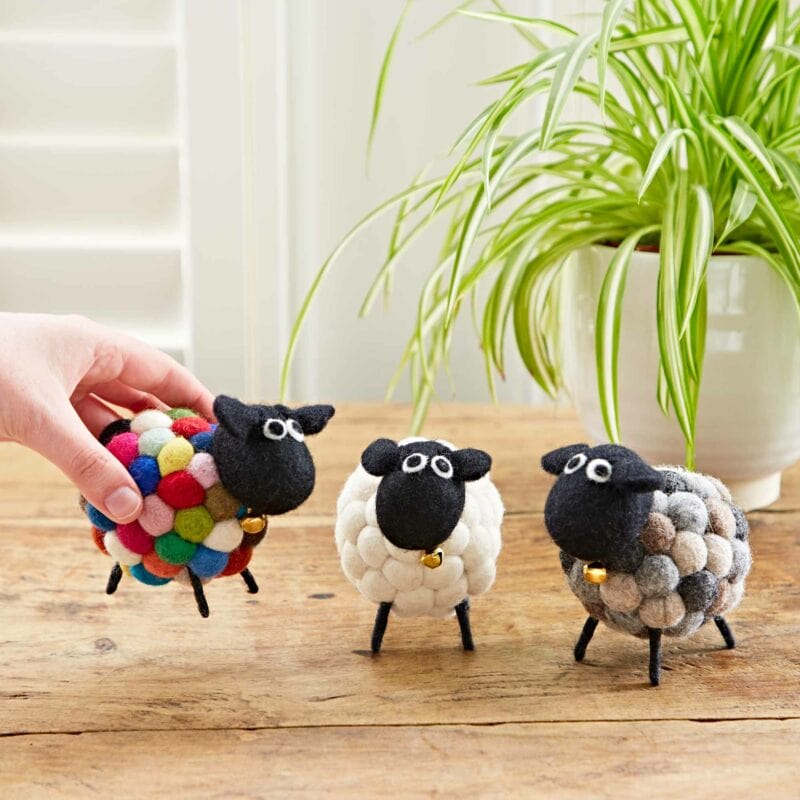 A hand arranging three Felt Sheep Ornaments, made of colorful wool balls, on a wooden table, with a green potted plant in the background.