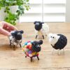 Four Felt Sheep Ornaments sit on a wooden table—three are black and white, one is colorful. A hand adjusts a felt sheep figurine while a potted plant appears in the background.