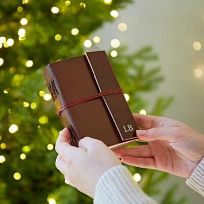 A person holding a brown leather scrapbooking journal in front of a Christmas tree.