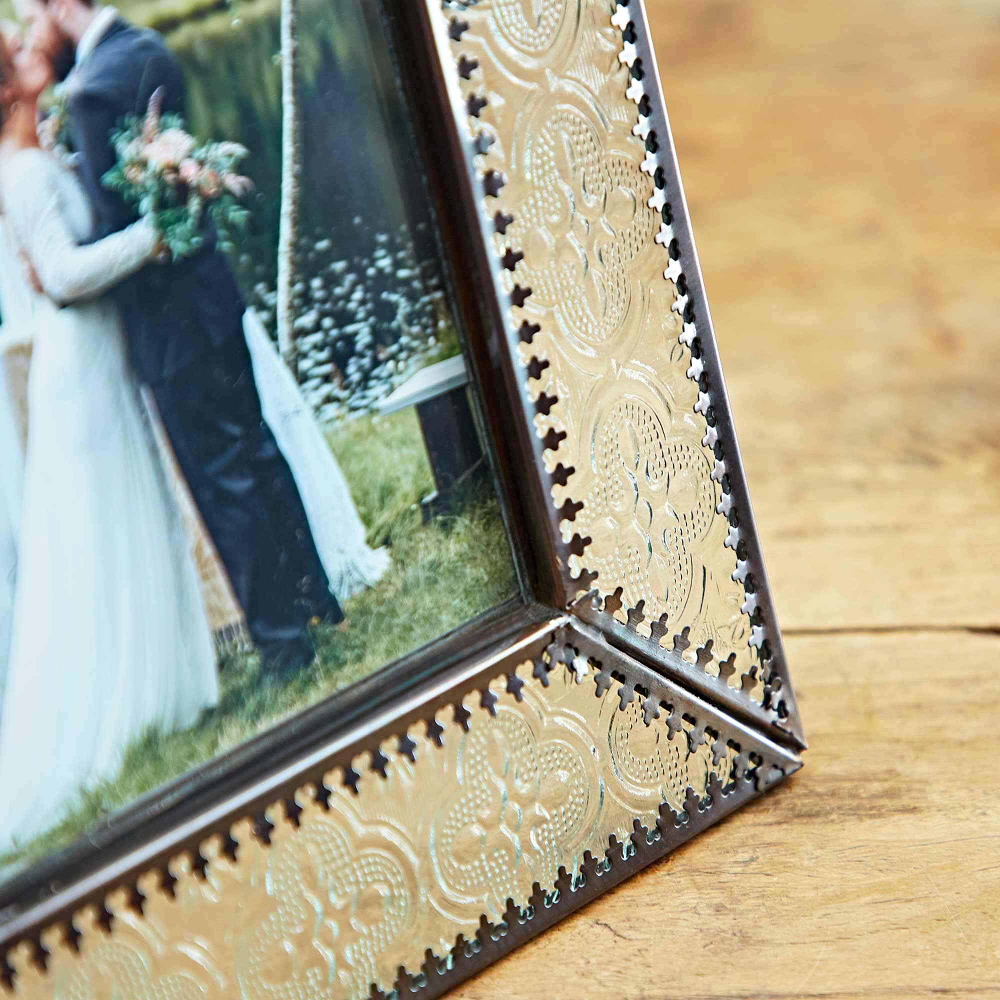 A close-up of a decorative 6" x 4" Luna Recycled Glass and Antique Brass Metal Photo Frame featuring an etched design. The photo within, nestled in this recycled glass photo frame, shows a standing couple embracing outdoors.
