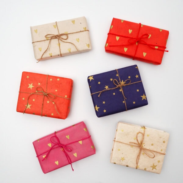 Six Lokta Paper Gift Wrapping Service boxes in red, cerise, blue, and natural colours with star and heart patterns, tied with jute strings, arranged on a white background.