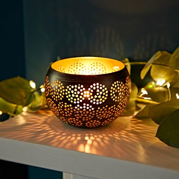 A Black and Gold Metal Tea Light Holder featuring a lit candle casts intricate patterns on a nearby surface, complemented by string lights and green foliage.