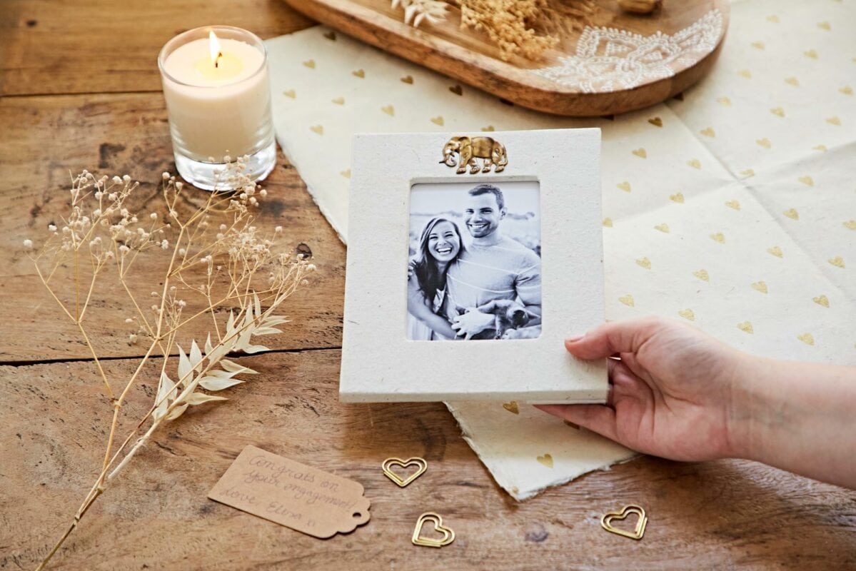 A hand holds a framed black-and-white photo of a smiling couple on a decorated table, ideal as an engagement gift. The scene is warmly lit by a candle, surrounded by dry flowers and heart-shaped ornaments.