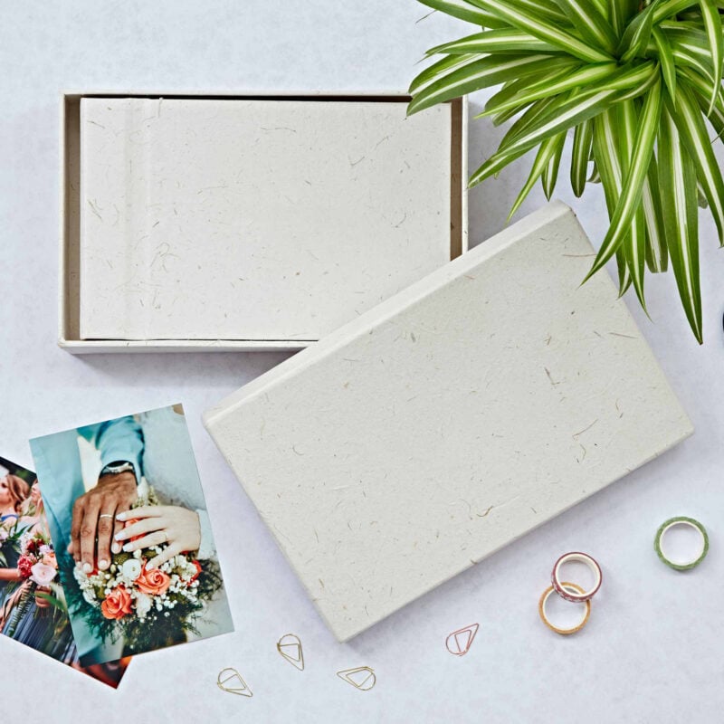 An open box with a blank card inside, surrounded by a plant, wedding photo, rings, and paper clips on a light background—perfect for curating thoughtful engagement gifts.