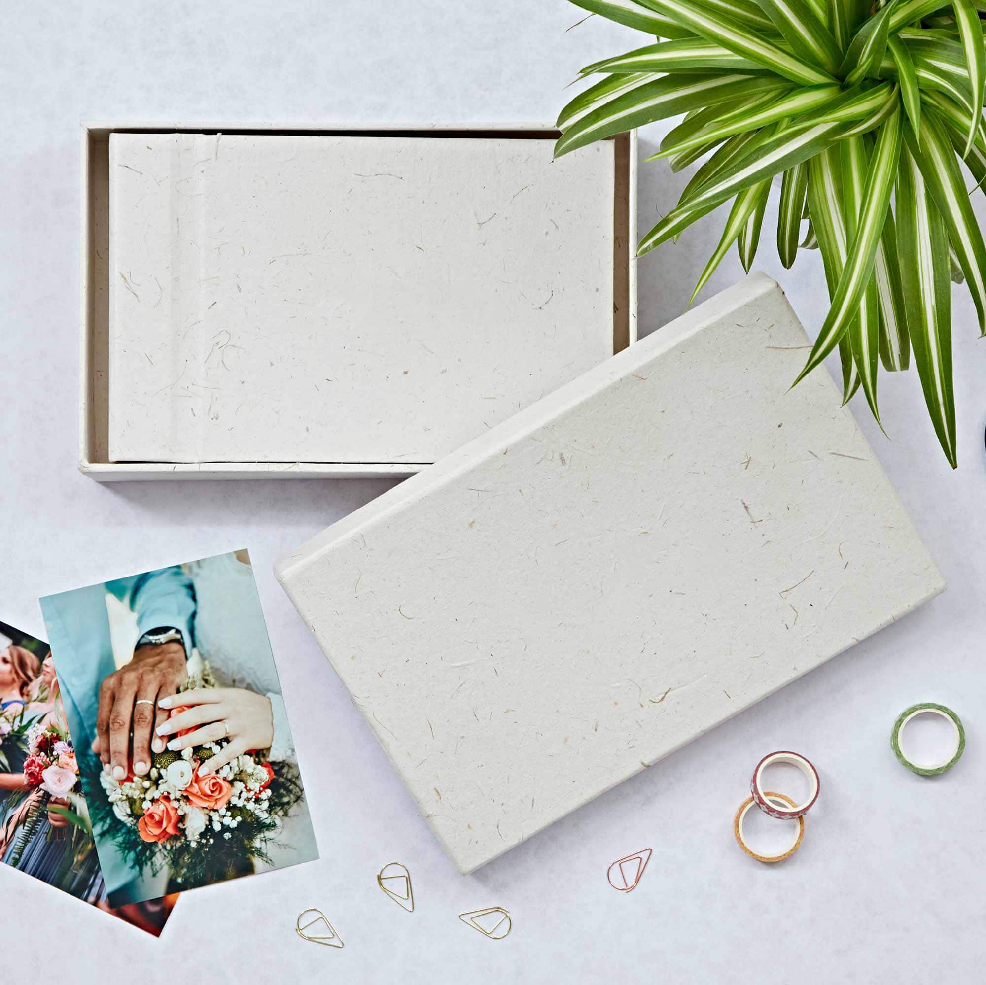 An open box with a blank card inside, surrounded by a plant, wedding photo, rings, and paper clips on a light background—perfect for curating thoughtful engagement gifts.