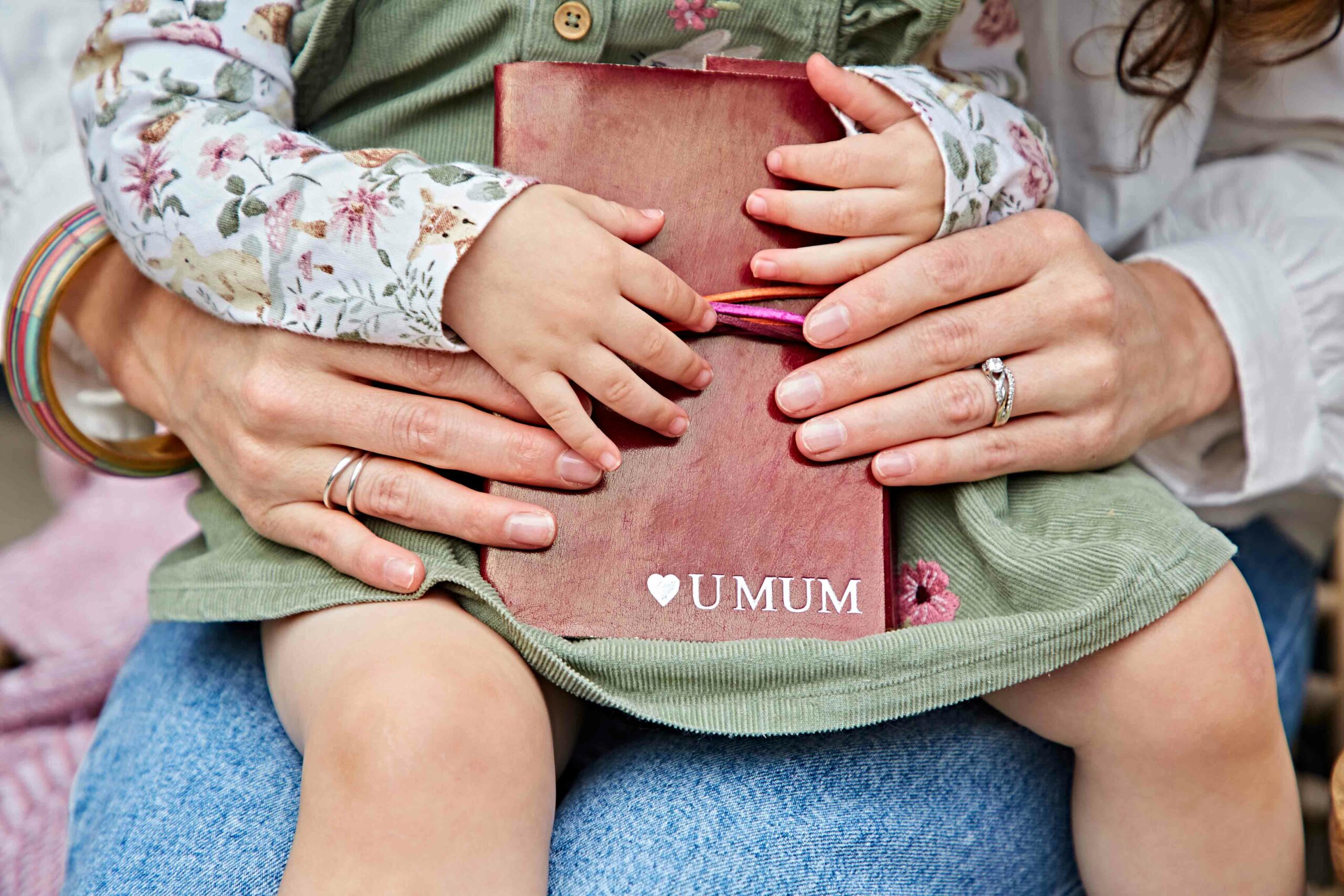 Adult and child hands holding a book with "UMUM" on the cover; child sits on adult’s lap, both wearing rings. A touching moment perfect for the Fairtrade Mother’s Day Gift Guide.