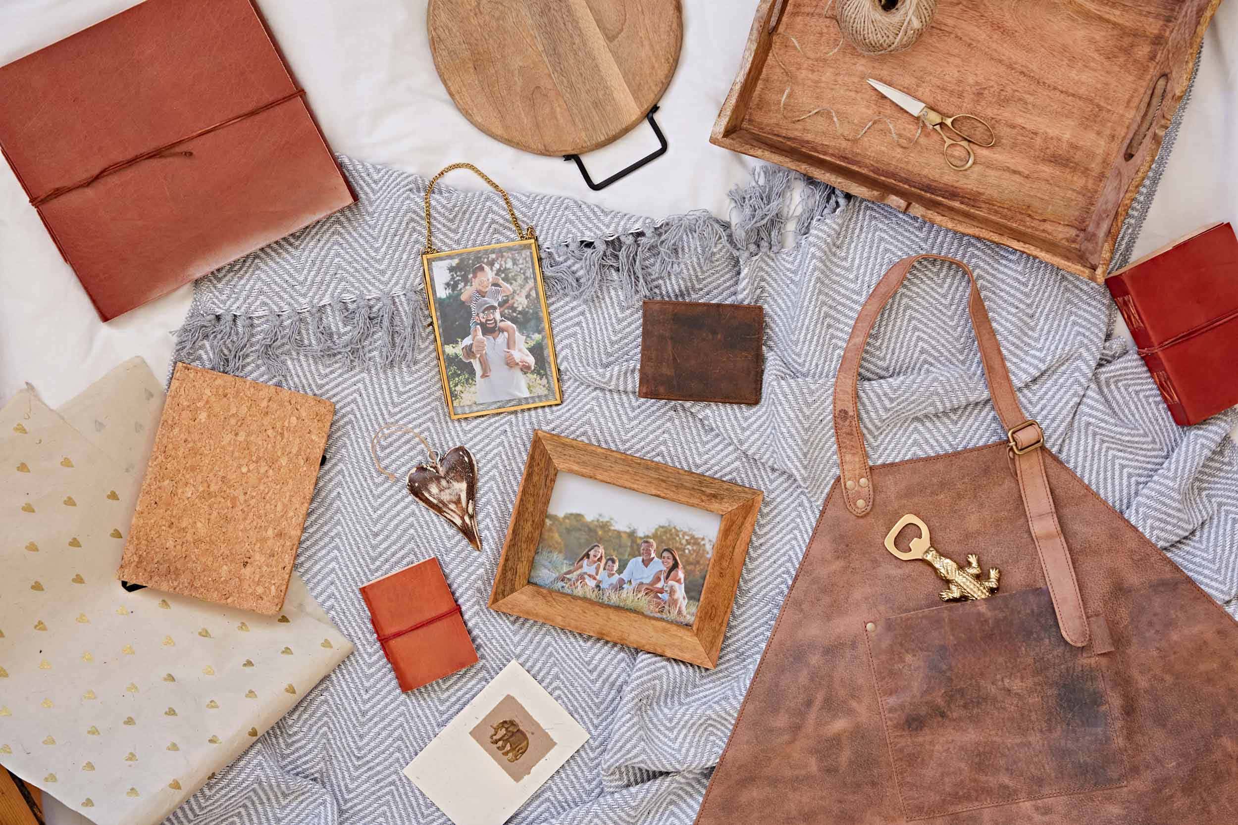 A flat lay of leather goods, framed photos, a wooden tray, scissors, wrapping paper, a cork notebook, and an apron arranged on a woven blanket for the Fair Trade Father's Day Gift Guide.