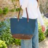 A person in a white long-sleeve shirt and blue jeans stands outdoors by greenery, holding a Personalised Buffalo Leather and Blue Canvas Tote Bag featuring the name "JANE.