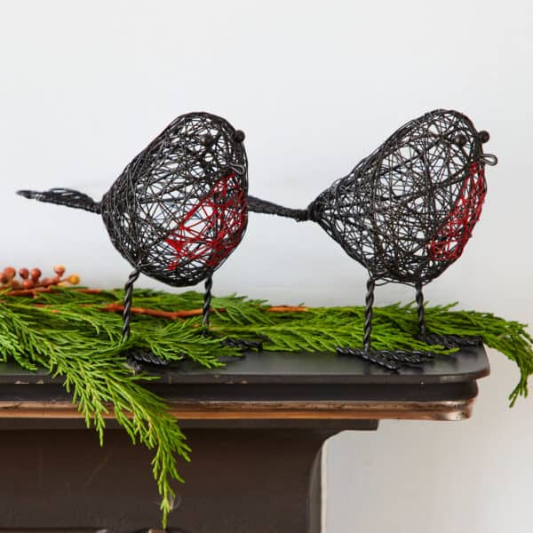 A Set of Two Wire Robin Decorative Ornaments with red accents stands on a mantelpiece decorated with green foliage and berries, set against a white background.