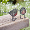 The Set of Two Wire Robin Decorative Ornaments, featuring red-accented chests, is displayed on a wooden bench outdoors amidst greenery and white flowers in the background.