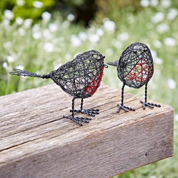 The Set of Two Wire Robin Decorative Ornaments, featuring red-accented chests, is displayed on a wooden bench outdoors amidst greenery and white flowers in the background.