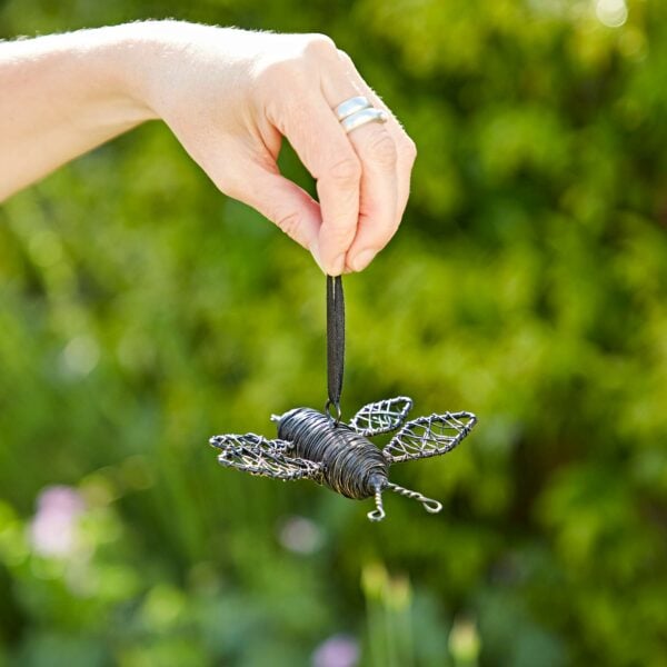 A hand holding a Hanging Garden Bee Ornament by a black ribbon with a blurred green background, perfect for adding charm to your hanging garden.