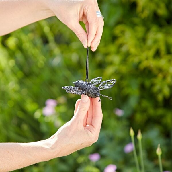 Two hands are holding a small, wire-crafted Hanging Garden Bee Ornament with a black ribbon against a background of green foliage, showcasing the perfect garden ornament.
