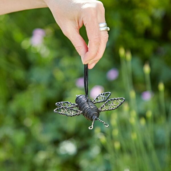 A hand with rings holds a metal wire bee ornament by a black ribbon, set against a blurred backdrop filled with green and flowers.