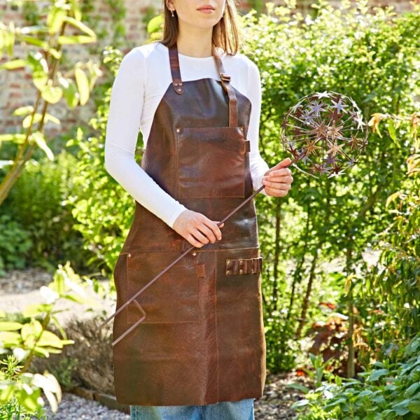 A person in a brown leather apron holds a Metal Allium Flower Garden Stake Decoration, standing in an outdoor garden setting.