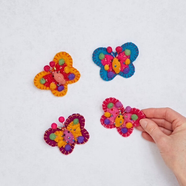 Four vibrant Felt Butterfly Hair Clips rest on a white background, with a hand elegantly picking one up. Each clip features intricate details, perfect for creating unique hairstyles.