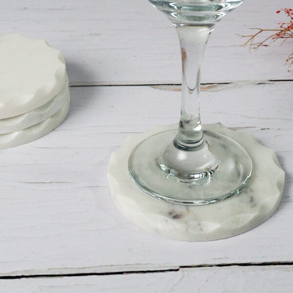 A glass sits on a round white marble coaster with scalloped edges, placed on a white wooden surface. Two matching coasters from the Set of 4 Round White Marble Coasters set are stacked nearby. Dried flowers partially appear in the top right corner, enhancing the elegant scene.