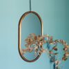 A Darpan Oval Antique Copper Hanging Chain Wall Mirror hangs on a chain against a light blue wall, with a branch of dried leaves partially reflected in the mirror.