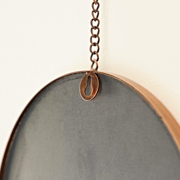 Close-up of a Darpan Oval Antique Copper Hanging Chain Wall Mirror hanging on a wall by a chain attached to a metal hook, with antique copper detailing.
