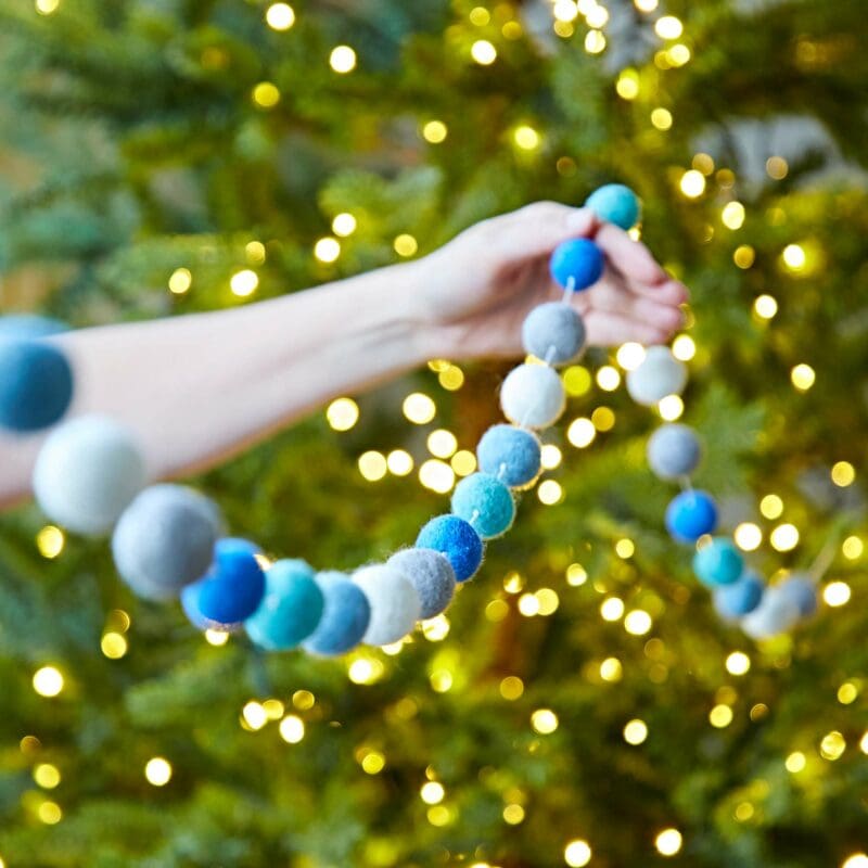 A person holding a Handmade Multicoloured Felt Ball Garland in front of a Christmas tree.