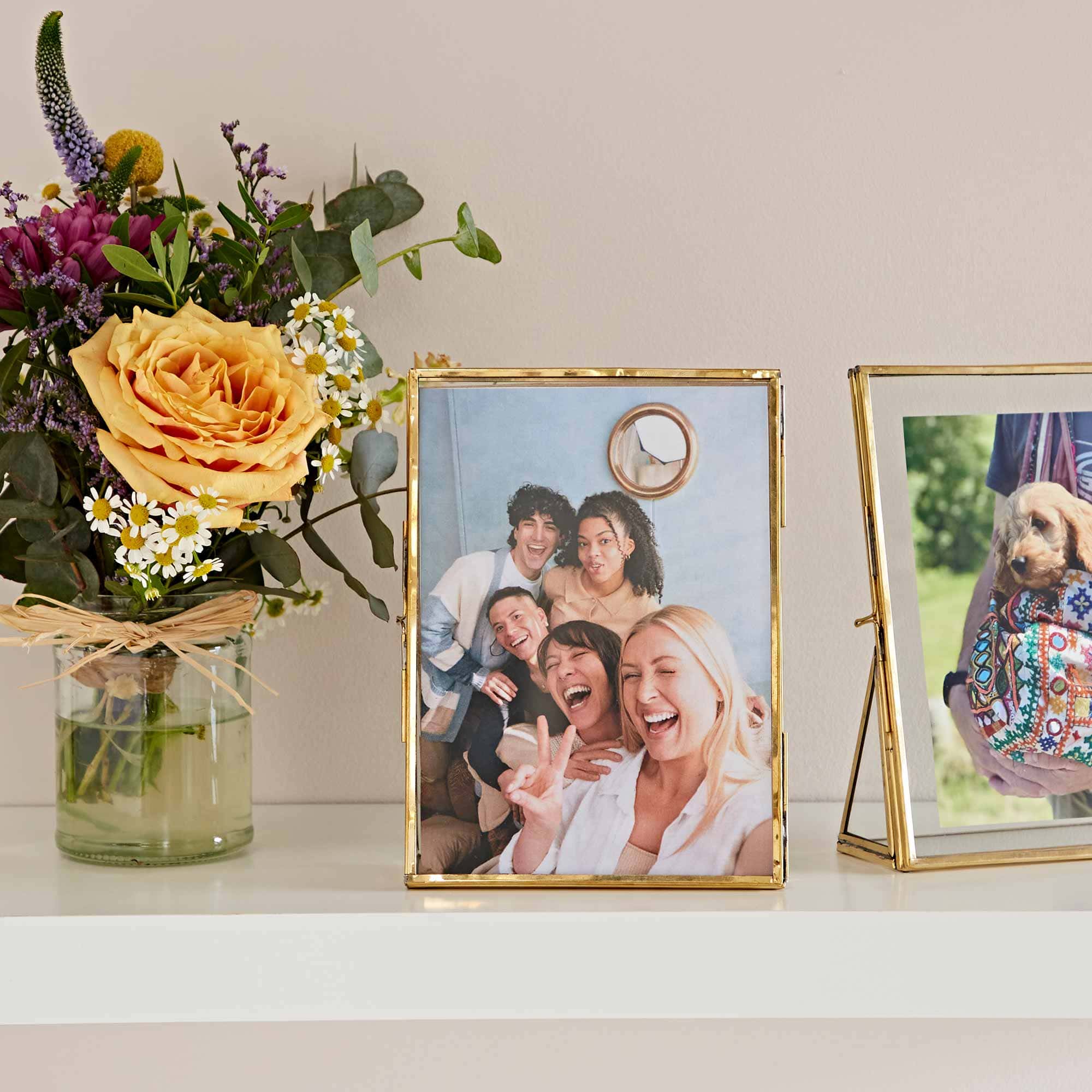 Two 7″ x 5″ Hiya Recycled Glass and Gold Metal Photo Frames and a vase of flowers on a shelf.