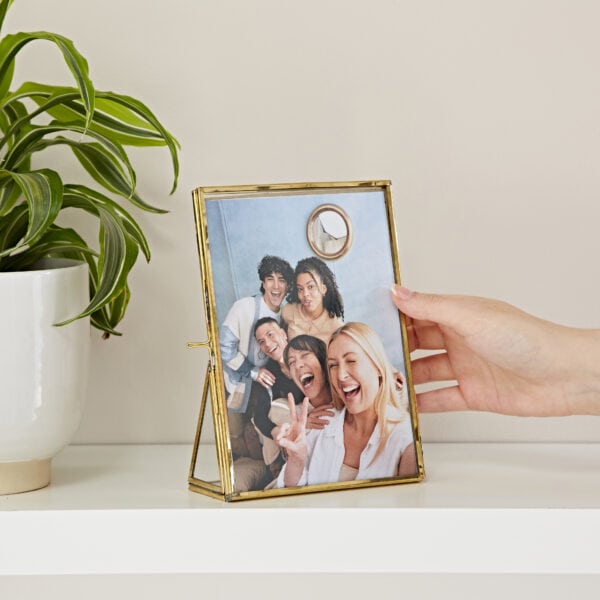 A person holding a 7" x 5" Hiya Recycled Glass and Gold Metal Photo Frame on a shelf.