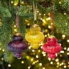 Three Anoli Coloured Recycled Glass Lantern Baubles in purple, yellow, and red hang from a Christmas tree branch, with warm white lights softly glowing in the background.