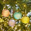 Four Ira Coloured Recycled Glass Ribbed Baubles in pink, blue, purple, and yellow hang from green tree branches, softly illuminated by blurry background lights.