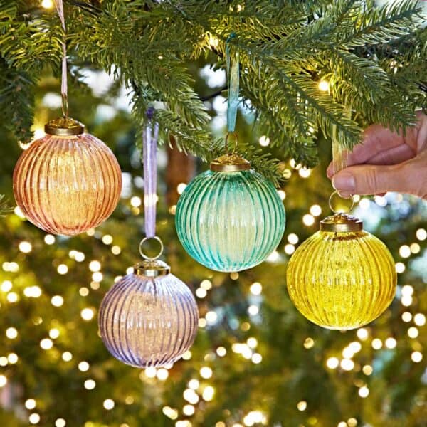 A hand hangs a yellow Ira Coloured Recycled Glass Ribbed Bauble on a Christmas tree, surrounded by pink, blue, and purple baubles, with fairy lights shining softly in the background.