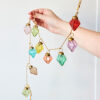 A hand holds the Mirai Pendent Coloured Recycled Glass Bauble Garland, featuring vibrant recycled glass ornaments in various shapes with red, pink, green, blue, and yellow hues against a plain background.