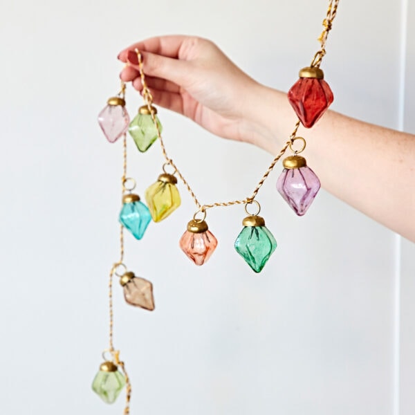 A hand holds the Mirai Pendent Coloured Recycled Glass Bauble Garland, featuring vibrant recycled glass ornaments in various shapes with red, pink, green, blue, and yellow hues against a plain background.
