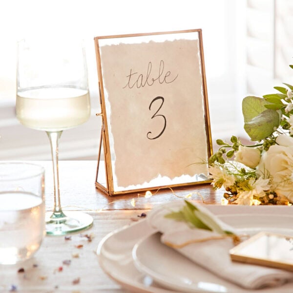Table setting with a sign that reads "Table 3," featuring a 7″ x 5″ Hiya Recycled Glass and Gold Metal Photo Frame, a wine glass, a water glass, and a plate with a napkin. Flowers and fairy lights are used for decoration.