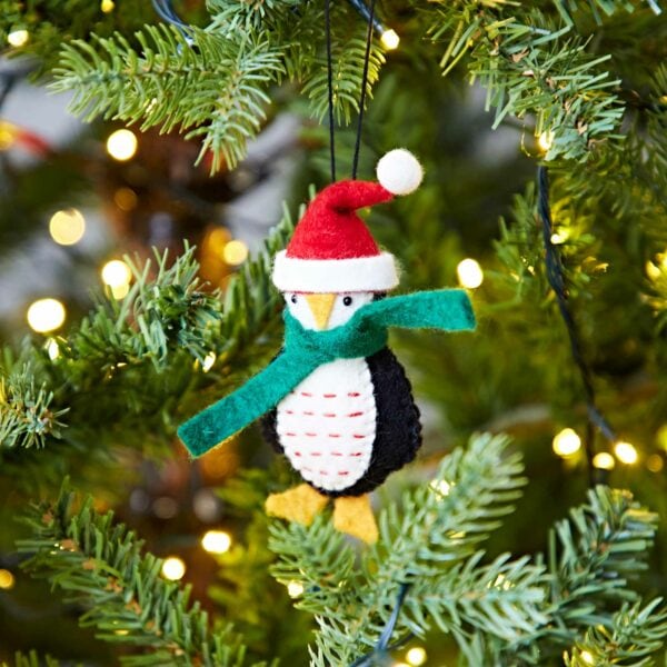 A Felt Penguin In Santa Hat Christmas Decoration hangs from a Christmas tree.