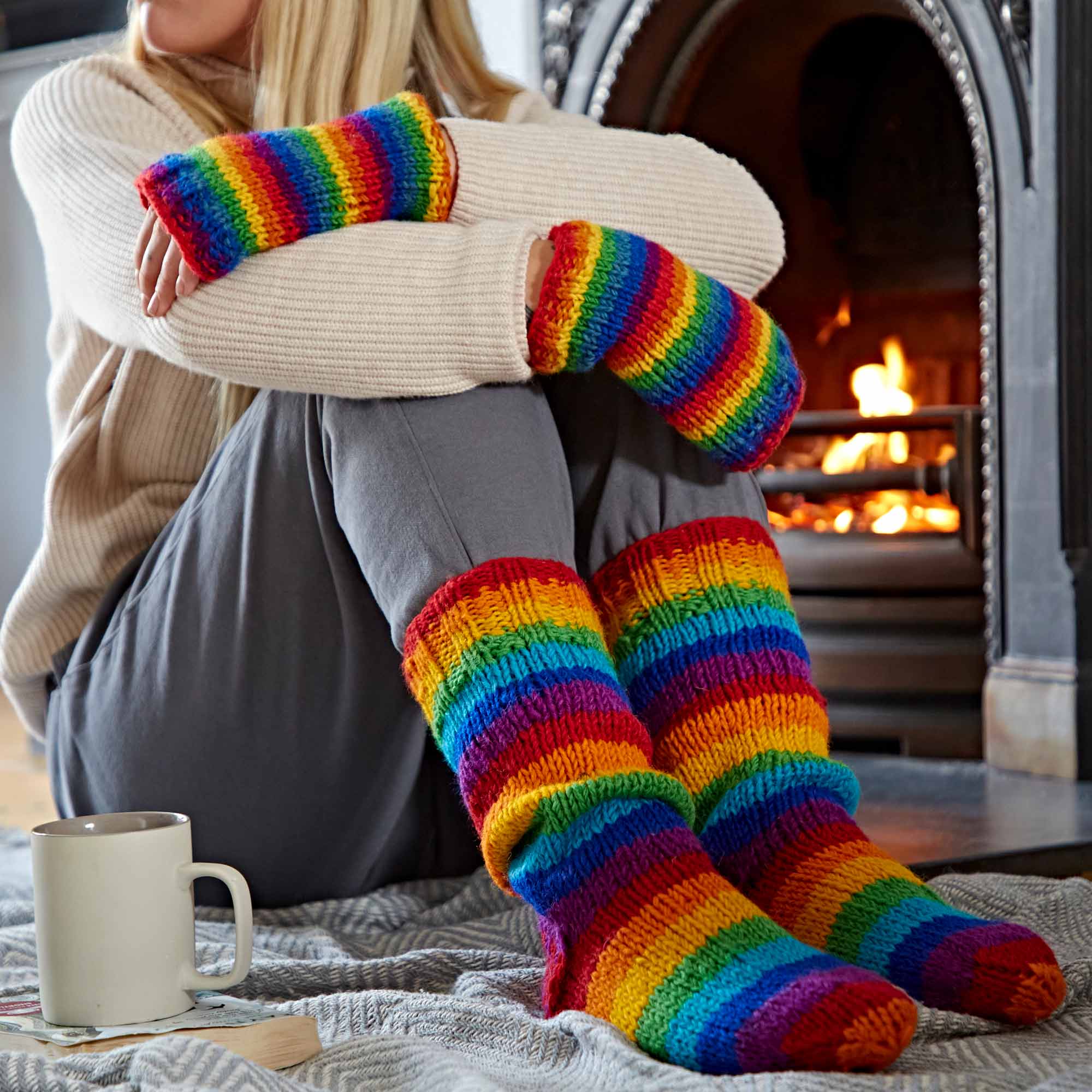 A woman is sitting on the floor with a cup of coffee and a Woollen Rainbow Handwarmer Gloves & Socks Set.