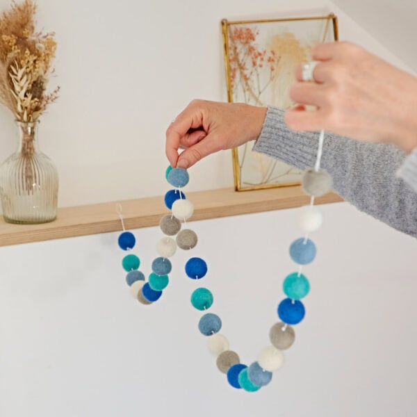 Person hanging a handmade felt ball garland of blue, white, and gray on a wall or shelf, with a vase of dried flowers and a framed artwork in the background.