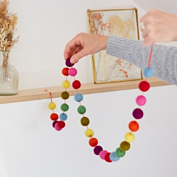 A person in a gray sweater is hanging a 1.3m handmade felt ball garland on a wooden shelf adorned with dried flowers in a glass vase. The multicoloured balls add a vibrant touch to the rustic decor.