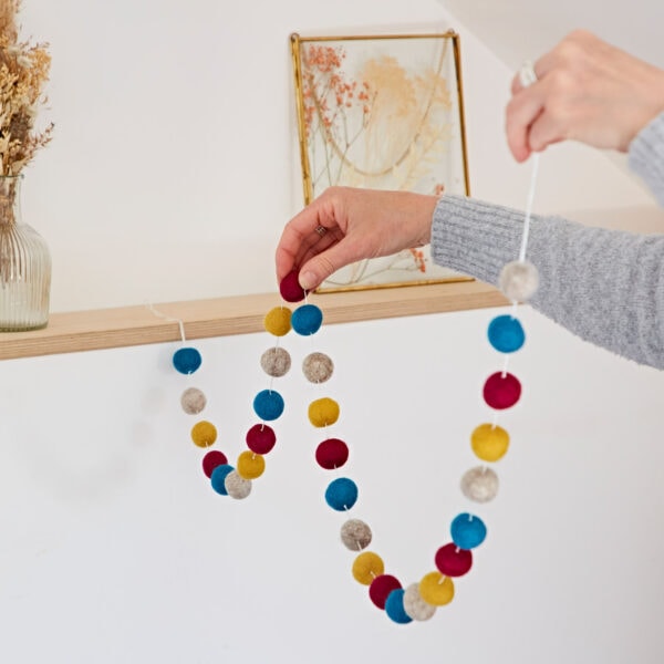 A person is hanging a 1.3m handmade felt ball garland on a wooden shelf, with a vase of dried flowers and framed artwork in the background.