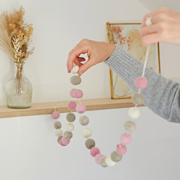 A person holding a handmade felt ball garland in white, pink, gray, and beige beside a wooden shelf with a vase of dried flowers and a framed picture in the background. The 1.3m garland adds a touch of charm to the cozy setting.