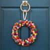 A Felt Multicoloured Wreath, vibrant in its woolen hues, adorns a blue door beside a silver knocker.