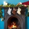 Three Felt Animal Christmas Stockings hang on a decorated fireplace mantel with candles and greenery. A hand places an orange object into the Felt Animal Christmas Stocking in the center.