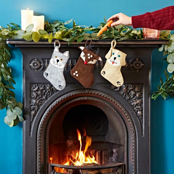Three Felt Animal Christmas Stockings hang on a decorated fireplace mantel with candles and greenery. A hand places an orange object into the Felt Animal Christmas Stocking in the center.