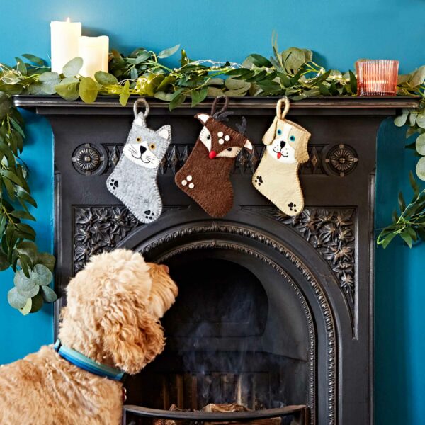 A dog looks at three Felt Animal Christmas Stockings hanging on a decorated fireplace, with lit candles and greenery above the mantle.