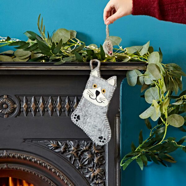 A hand places a Felt Animal Christmas Stocking, shaped like a gray cat, into a decorative black fireplace mantel adorned with green foliage.